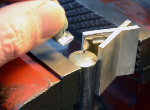The X shape is clamped in a vise. A steel dowel is positioned to force the X open enough to allow the stone to enter. Prior to this, the same part of the X shape was squeezed partly closed. This closing then opening of the shape results in just enough spring tension to lock the stone tightly in place.