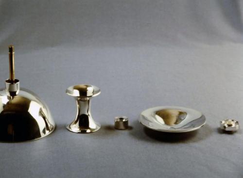 The finished Chalice disassembled. A threaded brass rod is soldered to the bottom of the bowl, and a sterling spacer is also soldered there. The flared stem will appear to hover in the design between this spacer and another spacer. The base fits on next, and the entire assembly is tightened together with a decorative threaded nut. This nut is made in a cross-like shape and is set with a central moonstone. All the surfaces have been given a mirror-like shine.