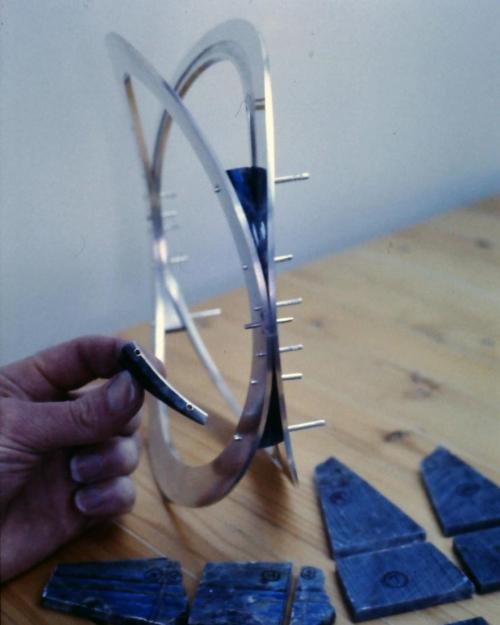 All the panels of labradorite have been cut from the slices. The four narrowest pieces have been finished. The piece in my hand shows how the stone has been backed with sterling sheet for strength (labradorite would otherwise cleave or break too easily). Onto the sterling back have been soldered two brass tubes (these appear as little dark-centered circles in the example I am holding). These tubes will receive the rivet wires. The labradorite is carefully fit to the sterling backing with its tubes, then is 